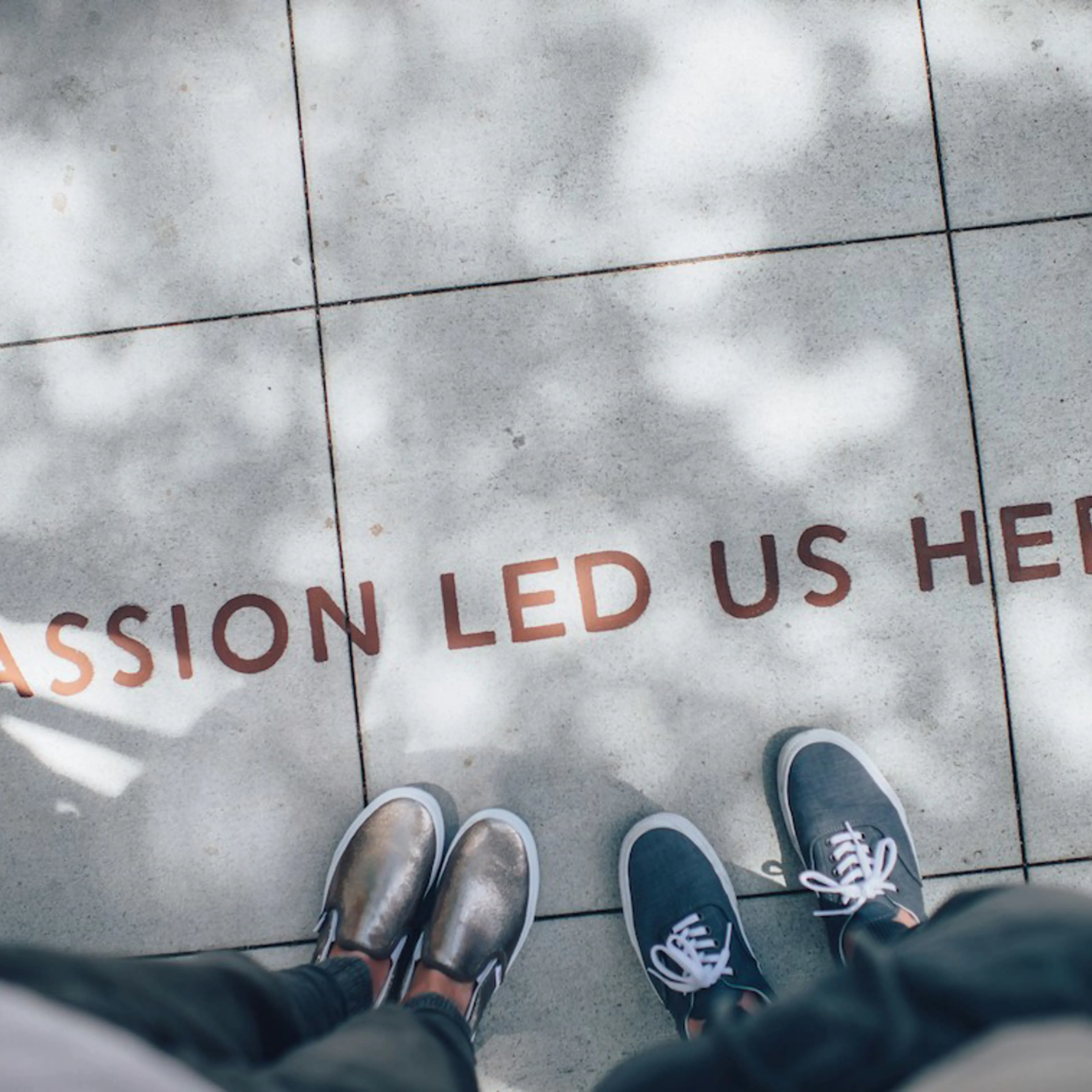 "Passion led us here" printed on floor tiles, and seen from above with 2 pairs of feet showing below it