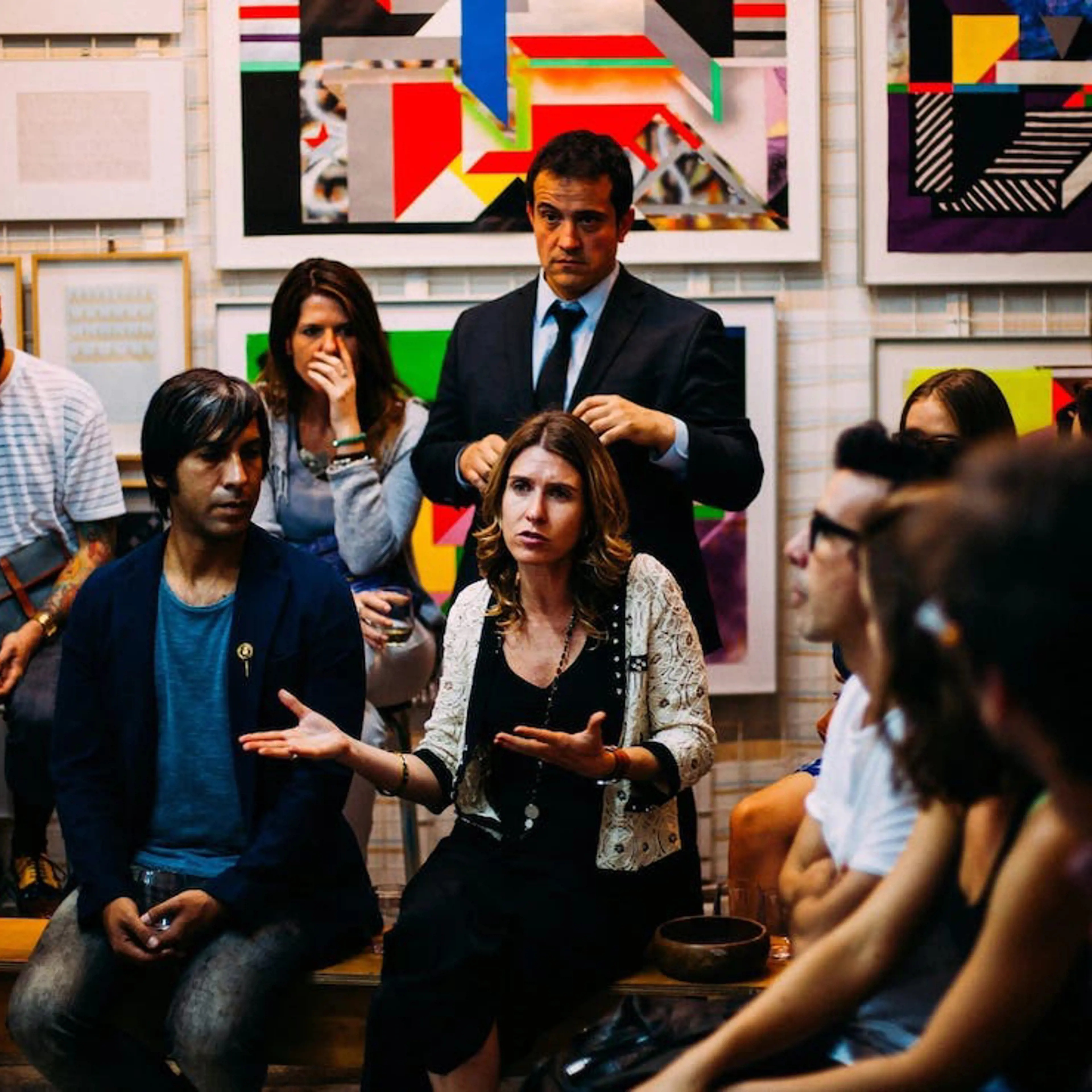 A lady talking among a group of people, some sitting, some standing, with art prints hanging on the background walls 